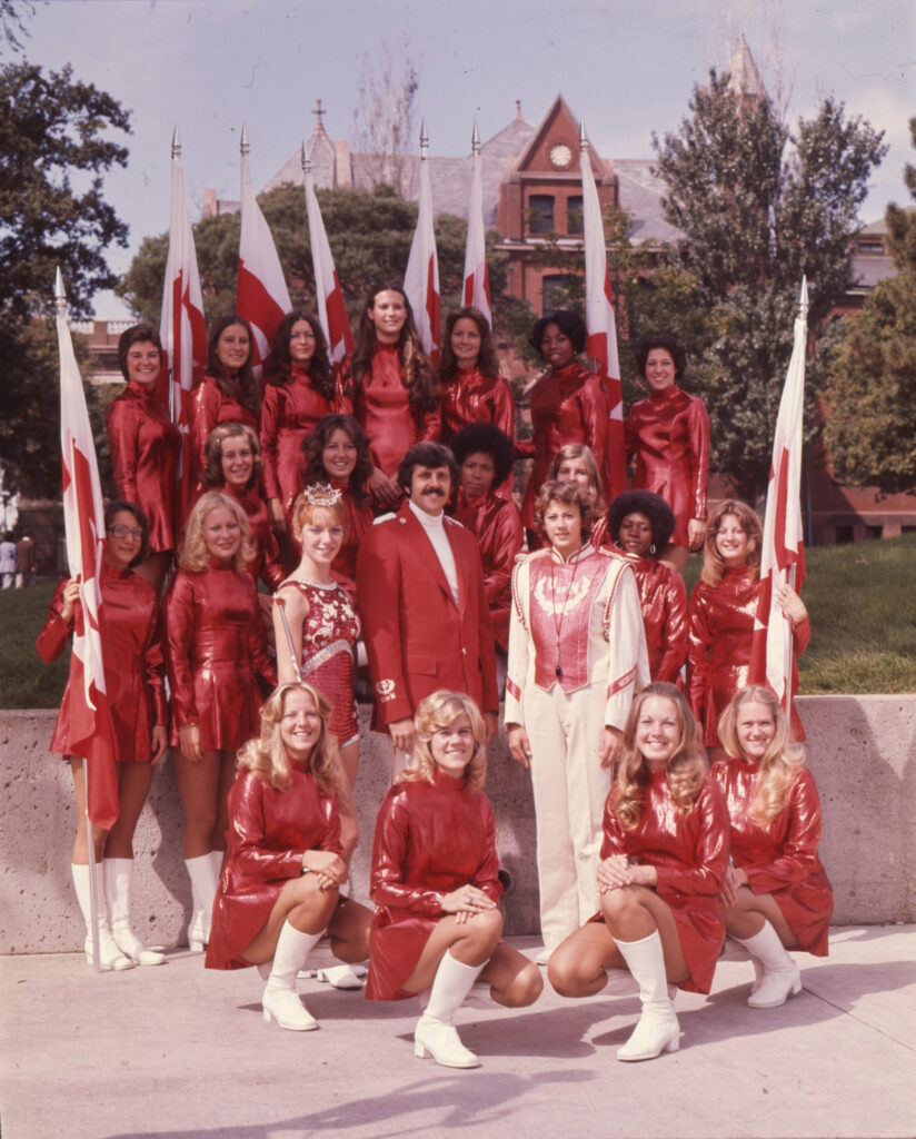 Women in red uniform with flags with a twirler, band director, and drum major.
