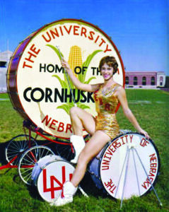 Baton twirler, Joyce Burns, sitting on drum with large drum behind.